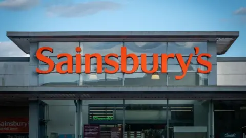 Entrance to a supermarket with a glass front and an orange Sainsbury's logo on top.