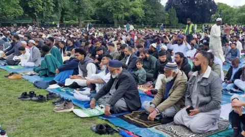 Crowds of men are sitting on prayer mats in a park, wearing thawbs. 