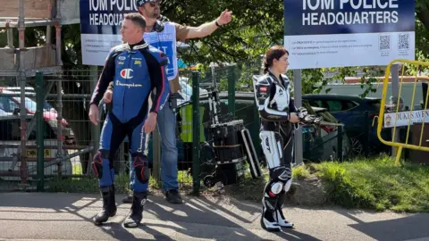 Frank Schuengel Channing Tatum is standing in blue and black leathers on a pavement on the left, with a crew member gesturing someone out of shot on the right. Next to Tatum on the right is Eve Hewson wearing black and white leathers, and is holding gloves.