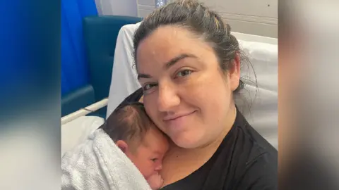 Close up image of Claire smiling whilst holding baby Osian. She is sat in a hospital bed. Clare has dark brown hair and is wearing a black t-shirt. Osian is wrapped in a towel and is resting on her chest. 