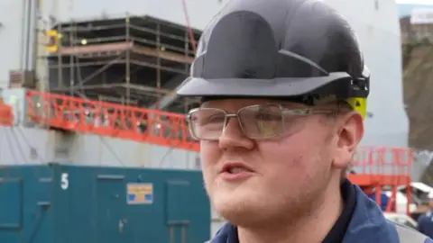 A worker wearing a black hard hat, safety glasses and a navy uniform stands in a busy dockyard with scaffolding and a large ship under repair in the background.
