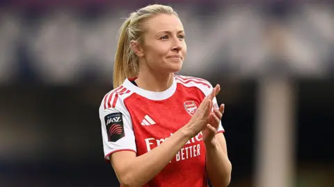 Leah Williamson, in a red Arsenal shirt with white sleeves and blonde hair tied back in a ponytail, looks in the direction of the crowd and applauds