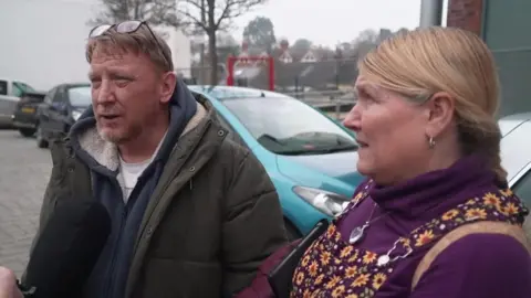 The image shows two people standing outdoors in what looks like a car park or a paved area with several parked cars behind them. One person is wearing a dark green padded coat with a hood and glasses, while the other is wearing a purple top with a patterned dress or pinafore and carrying a bag over one shoulder.