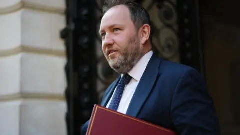 Ian Murray with dark hair and a greying beard walks in front of a stone building. He is wearing a blue suit and tie with a white shirt, and carrying a red folder. 