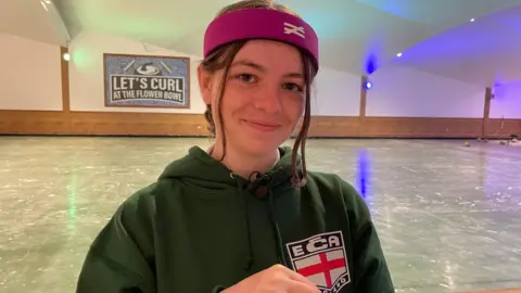 BBC Marinna smiles at the camera, wearing a cerise head band and a green England Curling Association hoodie, with the ECA logo featuring the English flag. The ice rink, with a sign saying 'Let's curl at the Flower Bowl' and an illustration of a curling weight and two brushes, can be seen behind her.