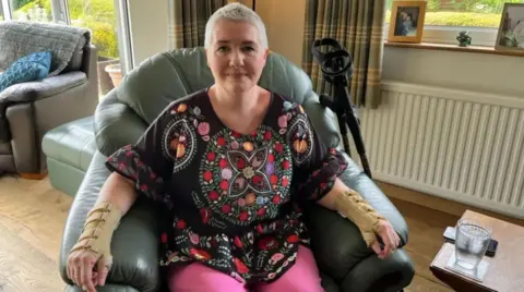 Angharad sitting in a leather armchair. She has cropped white hair and is wearing supports on her wrists and lower arms. She is wearing a black top with a colourful circular pattern on it, and bright pink trousers.