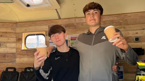 Corey (left) and Deacon (right), are standing inside a small kitchen in a converted horse box. They are both holding paper coffee cups. Both have short, brown hair. Corey is wearing a black top and Deacon is wearing a two-tone brown top.