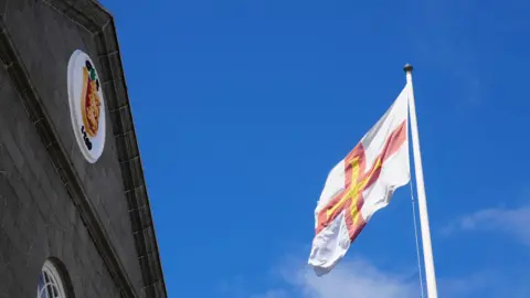 The top of the States of Guernsey chamber building. It is made of grey stone with an emblem of Guernsey on the front. A shield with three lions. In front of the building is a Guernsey flag flying on a pole. 