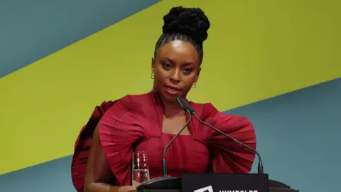 Chimamanda Ngozi Adichie, wearing a red shoulder fan dress, speaking into a microphone on a podium on stage, with a blue and green thick-striped wallpaper in her backdrop.
