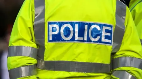 Getty Images A policeman is standing with their back towards the camera. They are wearing a yellow hi-vis jacket that says police on the back in white writing. 
