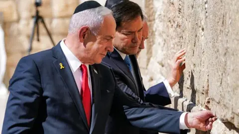 Getty Images Israel's Prime Minister Benjamin Netanyahu, US Secretary of State Marco Rubio, and US ambassador to Israel Mike Huckabee visit the Western Wall, Judaism's holiest prayer site, in the old city of Jerusalem. All three men are wearing suits and touching the wall. 