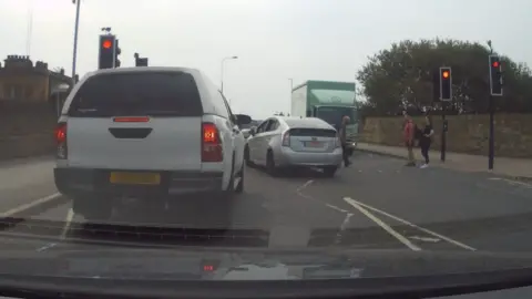 A van and a lorry stopped at traffic lights. A car has driven through a red light, and there are people crossing at the lights.