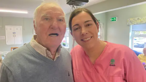 An older man with short white hair, wearing a white shirt and blue jumper. He is standing beside a woman with brown hair tied back, wearing pink scrubs.