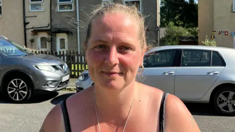 Head and shoulders shot of Deborah standing on the street n the sunshine. She has fair hair tied back and there are parked cars in the background