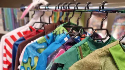 Nicola Rogers A metal rail with black wire hangers across it, holding various children's dress up costumes.