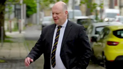 A bald white man in a dark suit and light shirt and dark tie walking up across a pavement by some cars