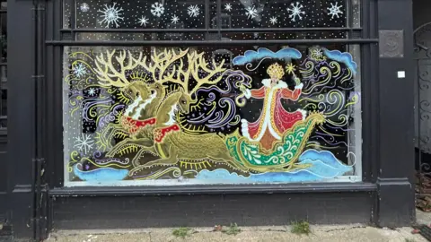 Art by Juliet Townsend A shop front has black paint and a large window. The window is painted with a figure riding a sleigh being pulled by ornately-decorated reindeer.