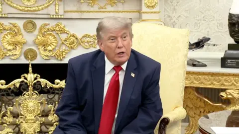Donald Trump sits in the Oval Office wearing a navy blue suit and red tie.
