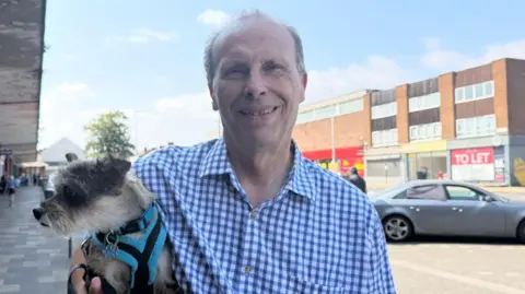 The photo shows Simon Shaw looking at the camera. He is wearing a blue checked shirt, and is holding a dog. He is standing in front of a high street of shops.