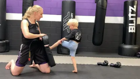 Two people are in the picture. The young blonde girl on the right is wearing light blue shorts and a black T shirt and is kicking a black pad with her left leg. The woman on the left of the picture is kneeling down, wearing a black gym top and purple shorts and is holding the black pad in front of her that the girl has kicked.