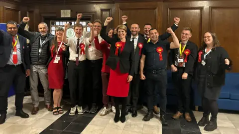 Charlie O'Loughlin/BBC MP Emma Hardy and canvassers wearing Labour Party rosettes cheer and smile for a photograph.