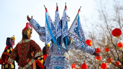 PA Media Stilt walkers in traditonal Chinese costumes, embroidered in intricate red and gold, and porcelain white and blue. Round red lanterns hang from tree branches in the background