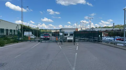 Tony Fisher/BBC A metal gate at the entrance to a Vauxhall factory with car parked in the background