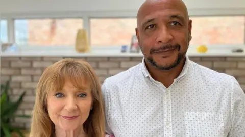 BBC Margo has shoulder length hair and is wearing a striped dress. She is stood next to her adult son, who is wearing a white shirt