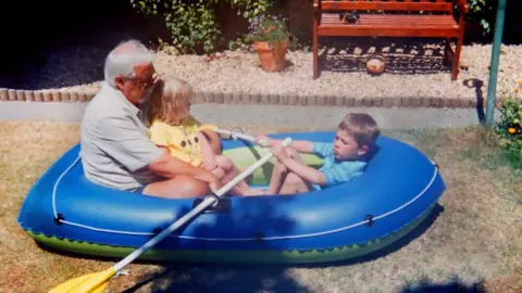 Ailsa Cleveland A picture of Trevor in a boat with his grandchildren. He is wearing a grey shirt. 