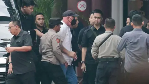 Getty Images is a man wearing a white shirt, a black hat and a black mask for the face, walking away from a car in a crowd 