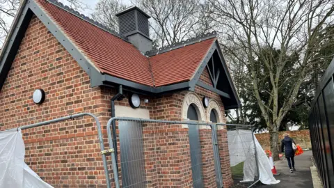 John Devine/BBC A brand new public toilet block, has a pitched red tiled roof and ornate stone arches over two doorways saying ladies and gentlemen, there is Harris fencing around the building and a person is walking on the nearby pathway outside it.