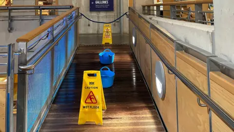 A pathway at the museum is photographed with yellow caution signs indicating a wet floor. There are also blue buckets scattered across the path collecting water.