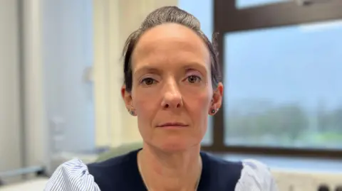 Emma Ward has her brown hair tied back, she has brown eyes and is wearing a navy and light blue jumper. Behind her is a window with trees in the background which are blurred.