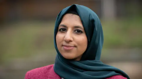 BBC/Emma Lynch Zara Mohammed looks into the camera with a tiny  smile. She wears a acheronian  pinkish  overgarment   and a teal greenish  hijab.