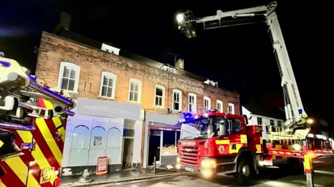 Fire engines are parked outside the a fish and chip shop where the glass 7in the windows on both floors is gone after fire damage. There is a large crane being used to access the damage. It is night time.