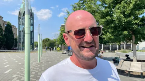 Laura Foster/BBC Jamie, wearing shades is standing in Basildon Town Centre. There are trees in the background and benches behind him.