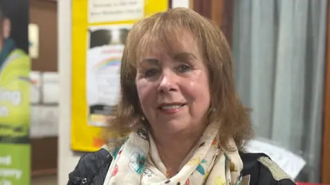 A woman with brown hair and a fringe. She wears a scarf with flowers on it. 