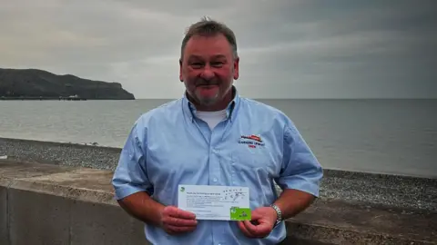 A man wearing a blue shirt stands holding a white letter against a sea short
