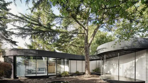 Royal Institute of British Architects A curved building with windows for walls go around a large tree. The roof of the building is black, otherwise the vertical windows go from floor to ceiling. There is a sofa in one of the rooms.