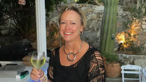 Contributed Hazel Jacobs, a woman sitting outside under a canopy. She is looking directly at the camera and smiling. She has blonde hair and is wearing a black crochet top. She is holding a glass in one hand.