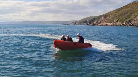 RNLI The west walk at Seaton, with the shore on the left-hand side of the image and a large path on the right-hand side with metal fencing separating the two. In the distance there are large white cliffs making up headlands and next to the path there are red cliffs with a bit of grass covering them.
