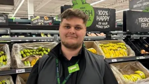Phil Snow-Griffiths has brown hair and a beard and is wearing an Asda lanyard and uniform. He stands in front of the fruit and veg section.
