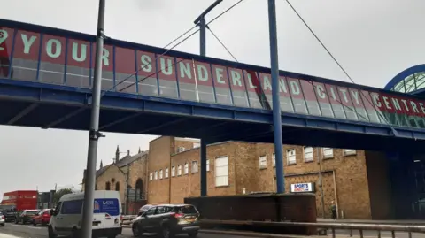 The bridge has a red banner saying Your Sunderland City Centre. It is above a brown brick Sports Direct building and there are cars passing underneath. The bridge itself looks to be enclosed and is made of grey and black metal with large rectangular window panes.