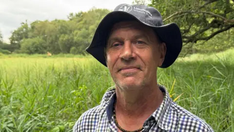 Phillip Norton/BBC Kevin James, a man wearing a rimmed hat and checked shirt, with a purple, white, blue and green pattern, smiles as he stands in a field of long, green grass, with green trees in the background.