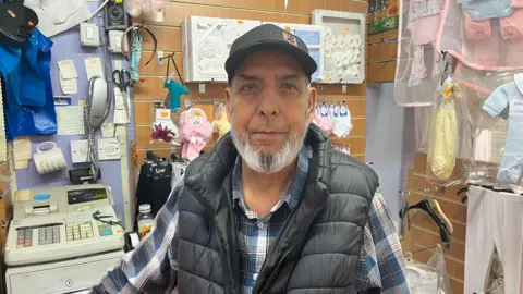 A man with a grey beard wearing a cap standing in front of a children's clothes shop