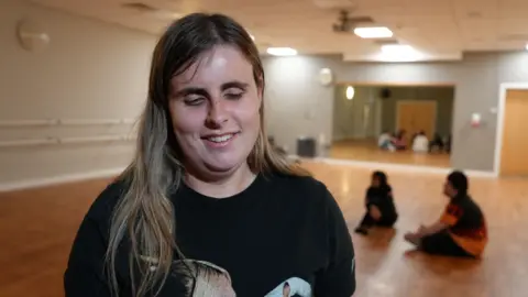 A girl with long fair hair smiles at the camera, whilst students sit behind her in a dance studio. She is wearing a black T-shirt with the top of a design on the front just visible at the bottom of the shot. The dance studio has a light wood floor, bars along one side and a mirror at the far end.