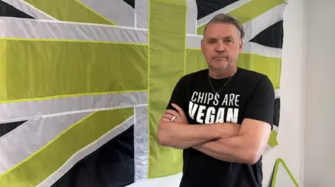 Dale Vince, in a black t-shirt with his arms crossed, stood to the right of the image in front of a green, white and black union flag.