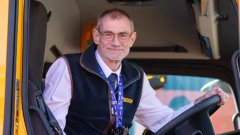 Gary Lawson - a gritter driver - sits in the front cab of a yellow gritter vehicle. He is wearing a white shirt and navy tie and a blue fleece with the yellow branding 'ECON' on it. Gary has short grey hair and wears silver rimmed glasses.