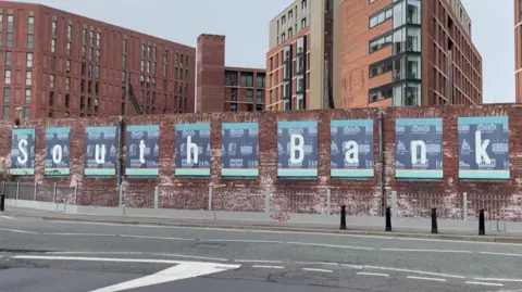 The words 'South Bank' are written in large white writing on top of blue boards stuck to a red brick wall. Multi-storey buildings are behind the wall, and there is an empty road in front of it.