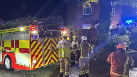 Bayliss family Four firefighters wearing protective uniforms and helmets are standing next to a fire engine - two of them are holding a hose. Behind them is a two-storey brick-built house.